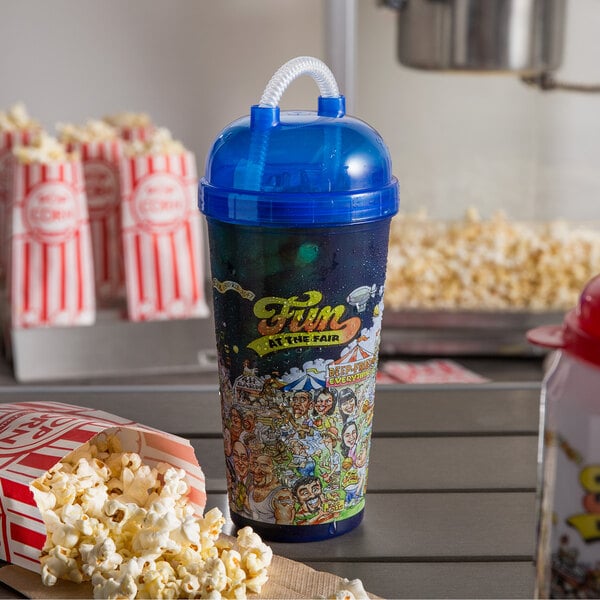 A blue "Fun at the Fair" Tizzeroo souvenir cup with a blue lid and straw next to a bag of popcorn.