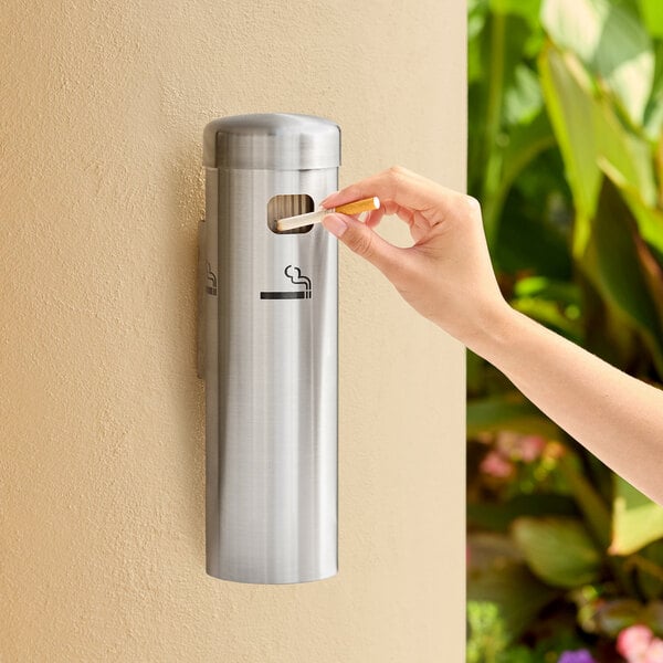 A stainless steel wall-mounted cigarette receptacle with a hand disposing of a cigarette.