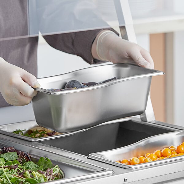 A person in gloves holding a Vollrath stainless steel container of food.