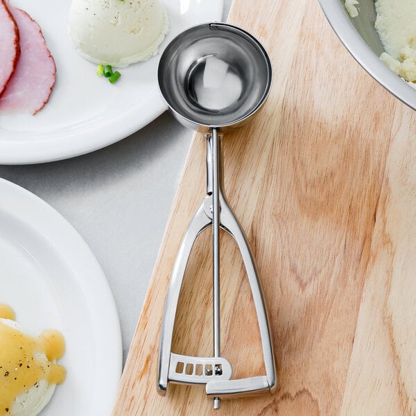 A stainless steel Vollrath round scoop with a metal squeeze handle on a wooden board.