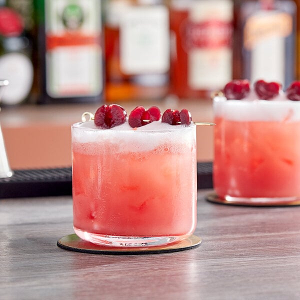 Two Acopa Spanish style double rocks glasses filled with pink drinks, ice, and cherries on a table in a cocktail bar.