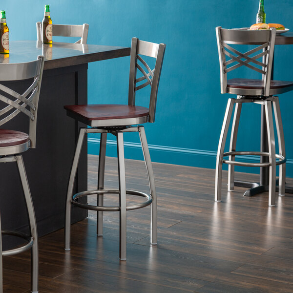 A group of Lancaster Table & Seating Clear Coat Finish Cross Back Swivel Bar Stools with Mahogany Wood Seats next to a table.