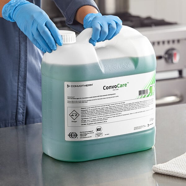 A person wearing blue gloves holding a large plastic container of green liquid.
