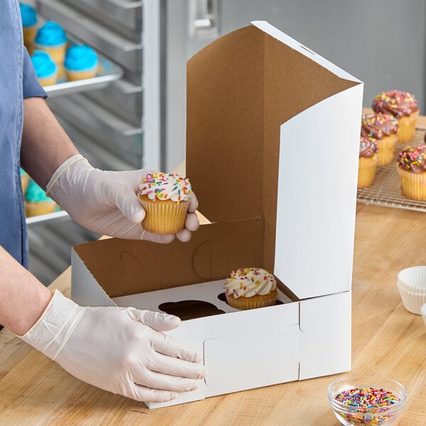 A white cupcake or muffin box with a reversible insert for holding six cupcakes, shown being filled with cupcakes topped with sprinkles.