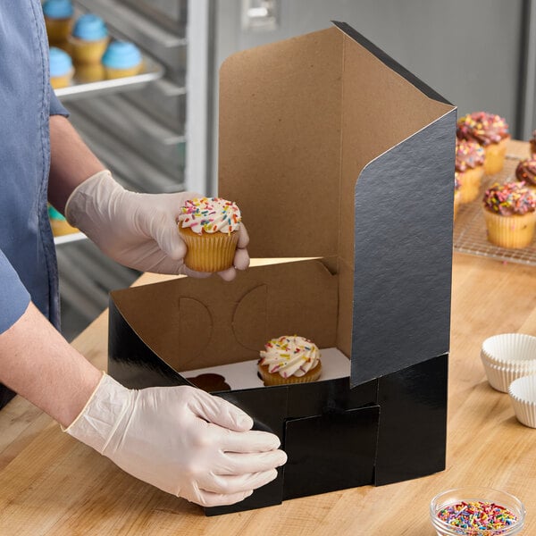 A black cupcake or muffin box with a reversible insert for holding six cupcakes, shown being filled with cupcakes topped with sprinkles.