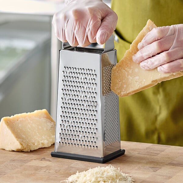 A four-sided stainless steel box grater with a soft grip handle being used to grate cheese.