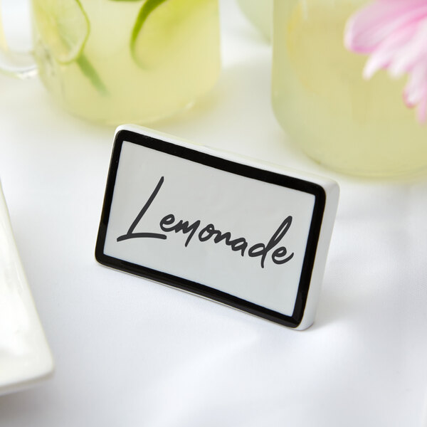 A black ceramic table tent sign with a black border on a table next to a white plate with a black border and a pink flower.