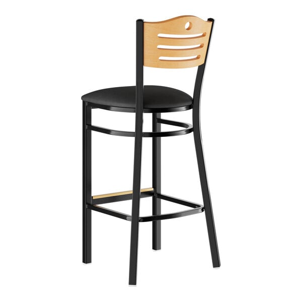 A bar stool with a black padded seat, black metal frame, and a light wood backrest featuring cut-out details.