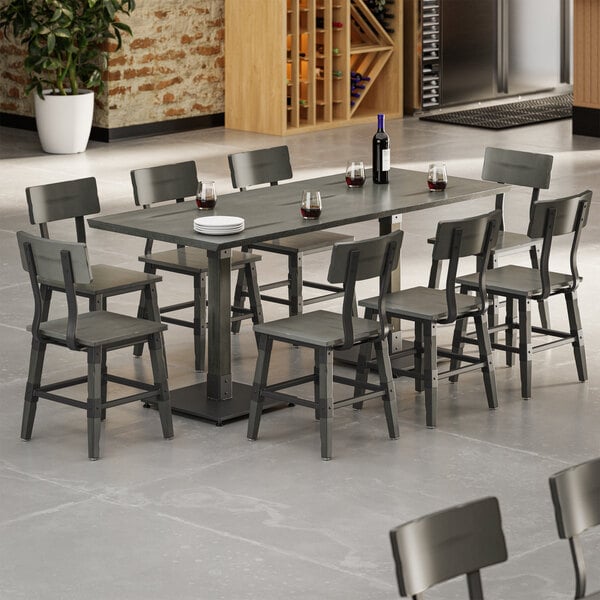 A rectangular dining table with a slate gray finish is surrounded by eight matching chairs, set with wine glasses, a wine bottle, and stacked plates in a modern, open space.