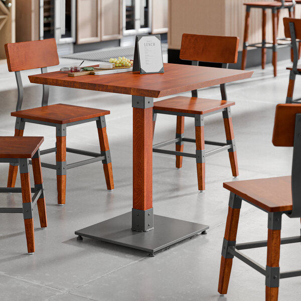 A square solid wood table with a rustic mahogany finish and industrial metal base, surrounded by matching chairs in a restaurant setting.