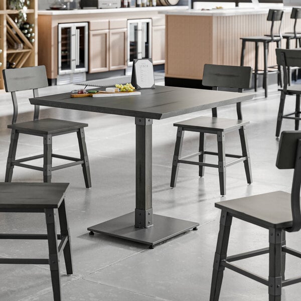 A square solid wood table with a slate gray finish and industrial design, surrounded by matching chairs in a modern dining area.