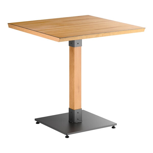 A square wooden table with a honey maple finish, supported by a single central wooden leg and a black metal square base.