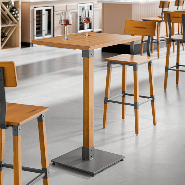 A Lancaster Table & Seating bar height table with two wooden stools and wine glasses on it.