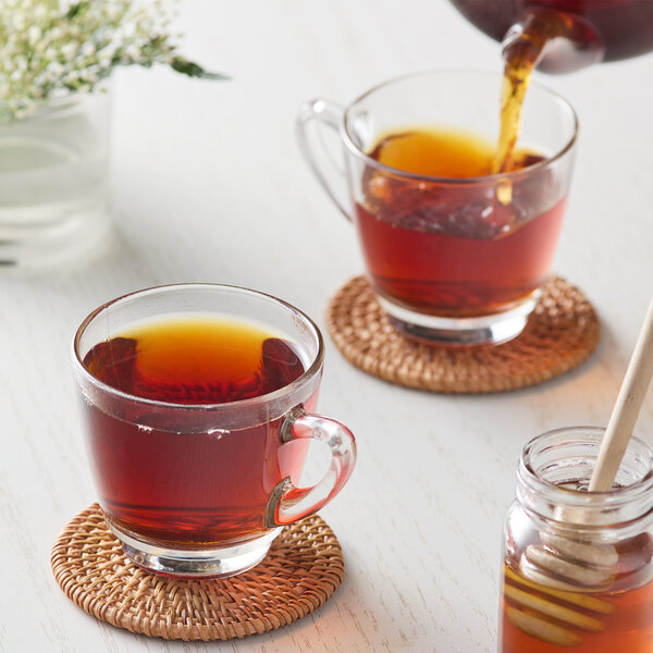 A glass cup of Numi Organic Chinese Breakfast Tea on a saucer with a jar of honey and a wooden spoon.