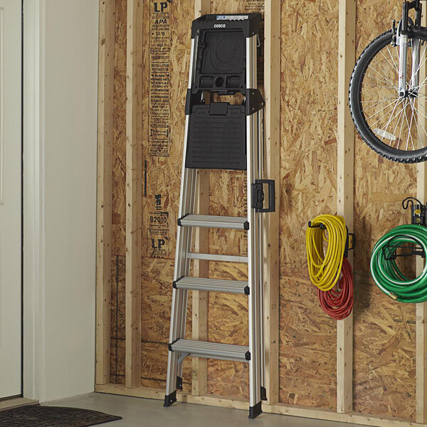 A Cosco aluminum step ladder with a work platform leaning against a wall.