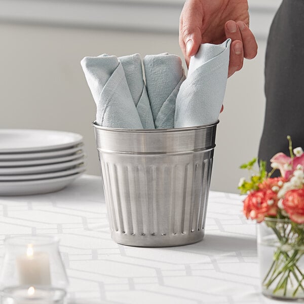 A hand holding a folded napkin in a round stainless steel utensil holder.