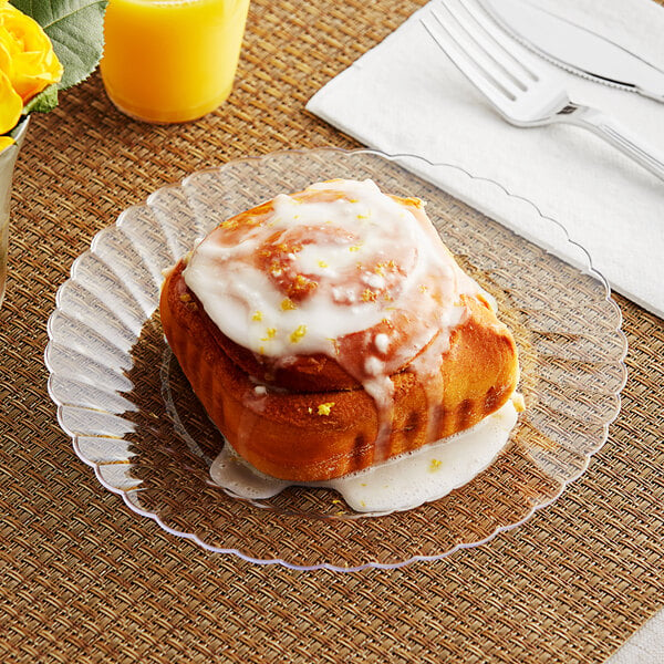 A WNA Comet Classicware clear plastic plate with a cinnamon roll on it next to a fork and knife and a glass of orange juice on a table.