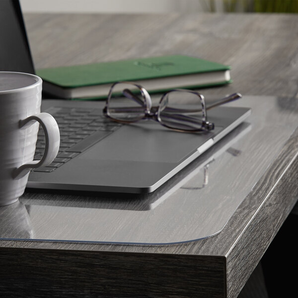 A clear ES Robbins desk pad on a table with a laptop and a white mug.