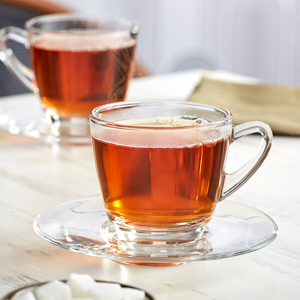 Two Acopa glass cups of tea on saucers with sugar cubes on a table.