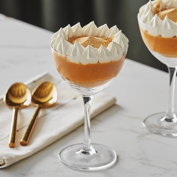 Two Acopa Nick and Nora glasses filled with dessert and topped with whipped cream on a table with golden spoons.