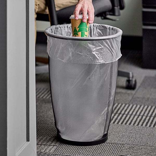 A hand putting a coffee cup in a Safco black round mesh wastebasket.