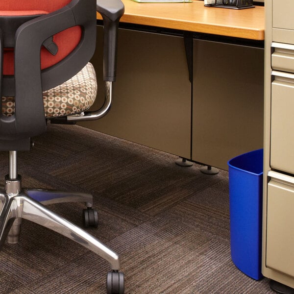 A blue Safco round wastebasket with a black border next to a black chair in a corporate office.