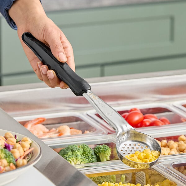 A hand holding a black Vollrath Spoodle over a tray of food.
