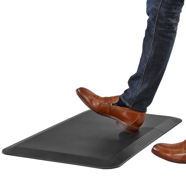 A person's shoe stepping on a black Safco anti-fatigue mat.