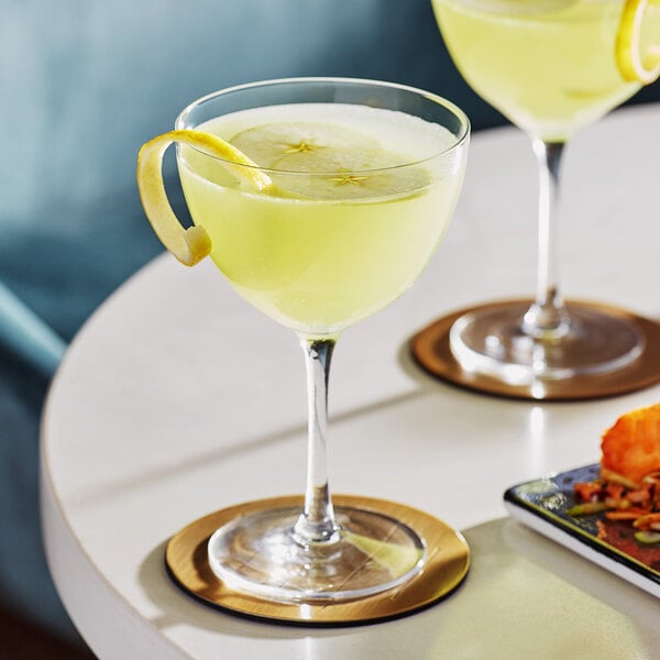Two Acopa Deco Nick and Nora cocktail glasses with yellow liquid and lemon wedges on a white table.