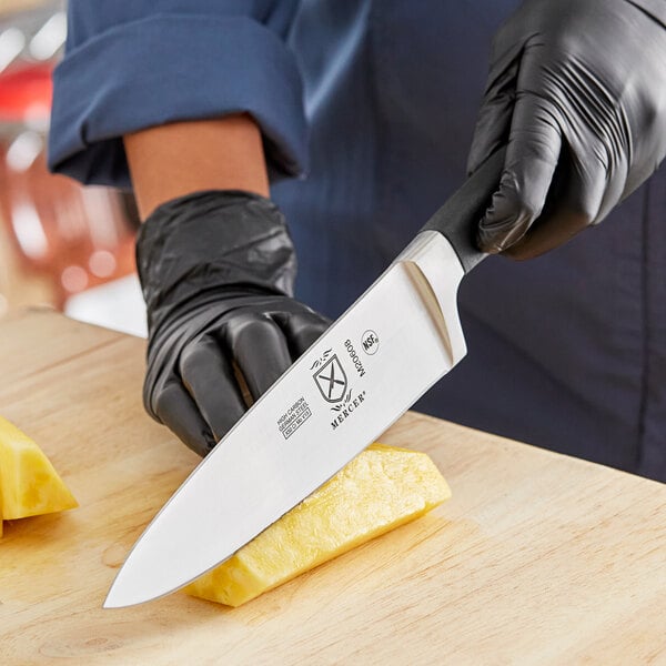 A person in black gloves using a Mercer Culinary Genesis chef knife to cut a pineapple on a cutting board.