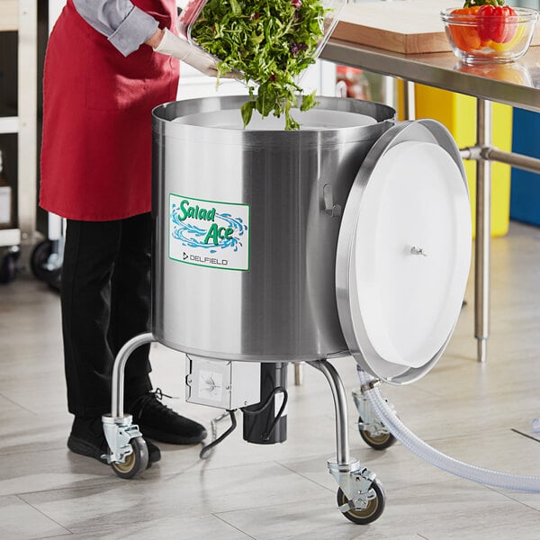 A person in a red apron using a Delfield salad dryer to dry green leaves in a large metal container.