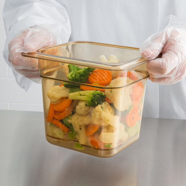 A person in gloves holding a Carlisle plastic container of vegetables.