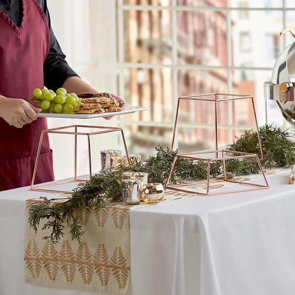 A woman using an Acopa rose gold metal display stand to serve food on a table with a white tablecloth and candles.