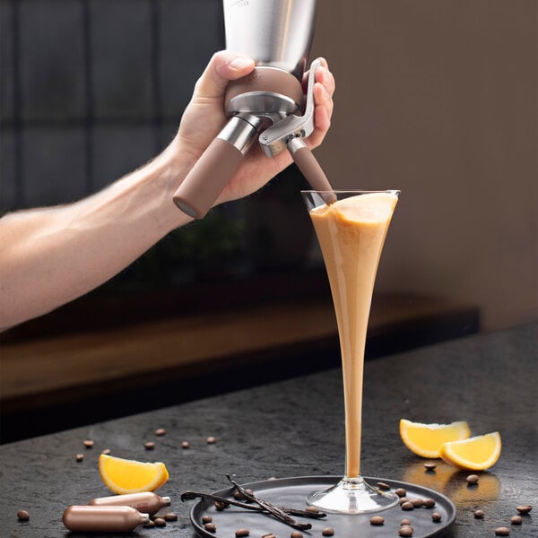 A hand is dispensing a creamy beverage from a stainless steel canister into a tall glass, with orange slices, chocolate pieces, vanilla beans, and coffee beans on the table nearby.