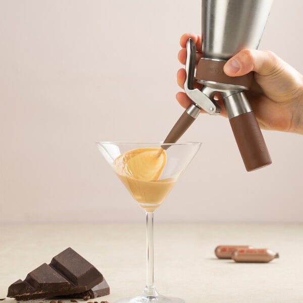 A hand is dispensing a creamy liquid from a stainless steel canister into a martini glass, with pieces of chocolate nearby.