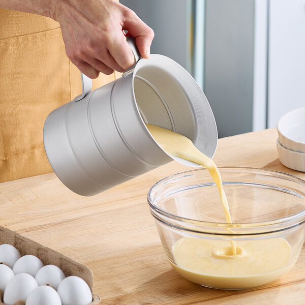 A hand pouring liquid from a Vollrath aluminum measuring cup into a bowl.