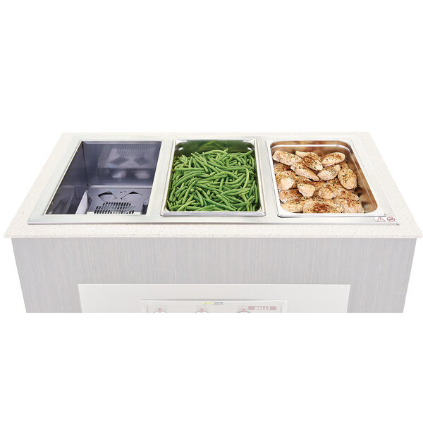 A white counter top with three Wells trays of food including green beans and chicken.