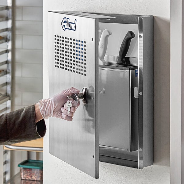 A hand in a white glove opening the door to a metal knife cabinet.