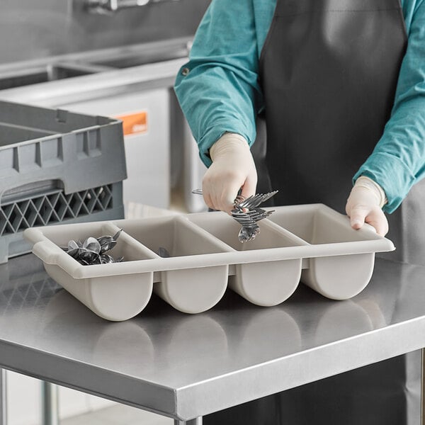 A person in an apron holding a gray Carlisle Save-All polyprolyene cutlery box with utensils inside.