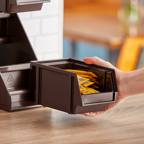 A hand holding a Vollrath brown condiment bin with packets of sugar in it.