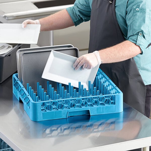A man in a blue apron putting a white plate into a blue Carlisle all purpose dish rack.