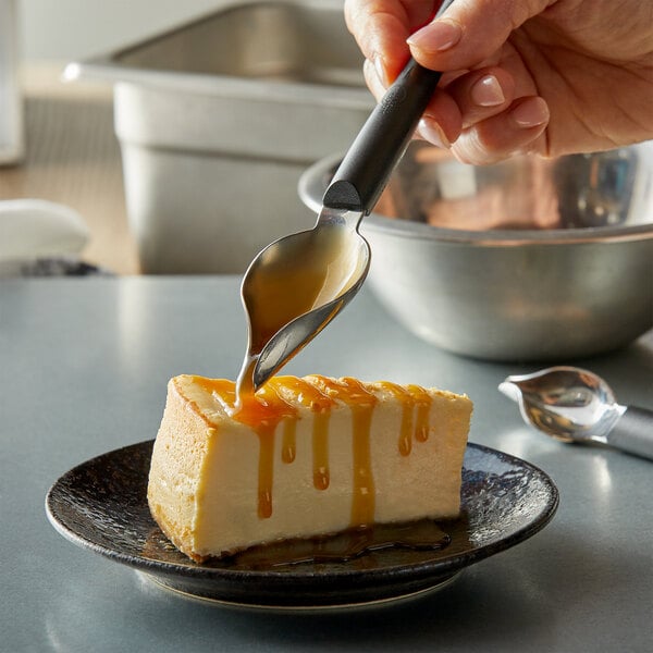 A hand holding a Mercer Culinary stainless steel precision spoon pouring caramel sauce over a slice of cheesecake.