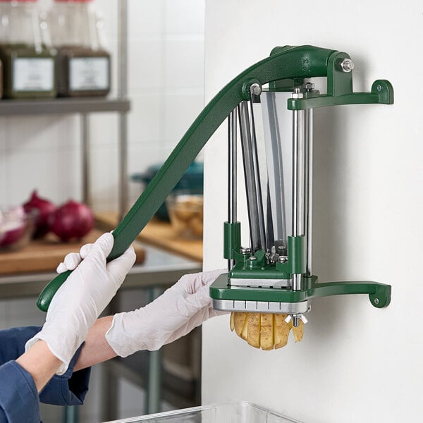 A wall-mounted green metal French fry cutter being used to slice a potato.