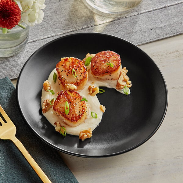 A Acopa matte black stoneware coupe plate with scallops and cream sauce with a fork on the table.