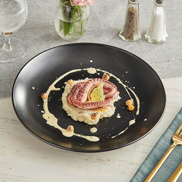 An Acopa matte black stoneware coupe plate on a table with food and a glass of lemon water.