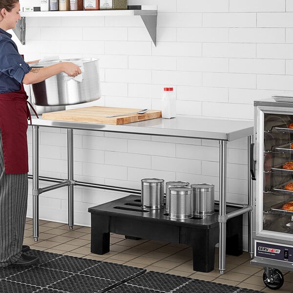 A woman in a red apron standing at a Regency stainless steel open base work table.