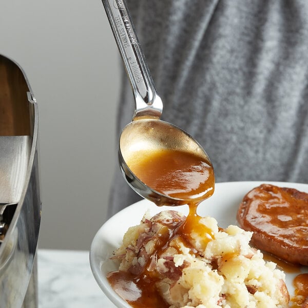 A person using a Vollrath black stainless steel spoodle to pour gravy on mashed potatoes.