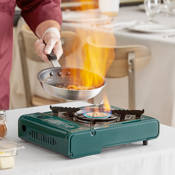A chef cooks food in a pan on a green Choice Green portable gas stove with a flame.