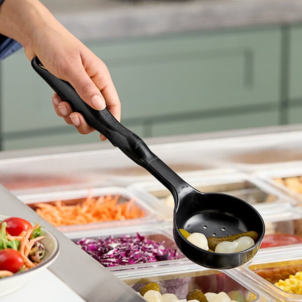 A hand holding a black Vollrath Spoodle over a bowl of vegetables on a salad bar counter.