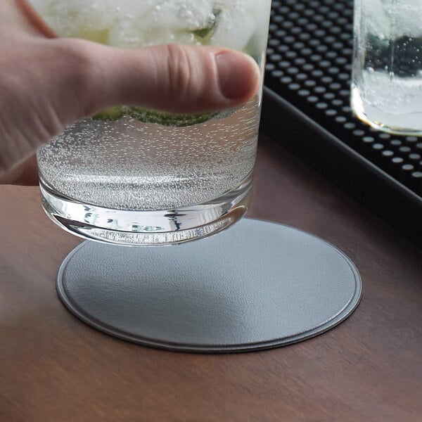 A hand holding a glass of water on a customizable silver vinyl coaster on a table.